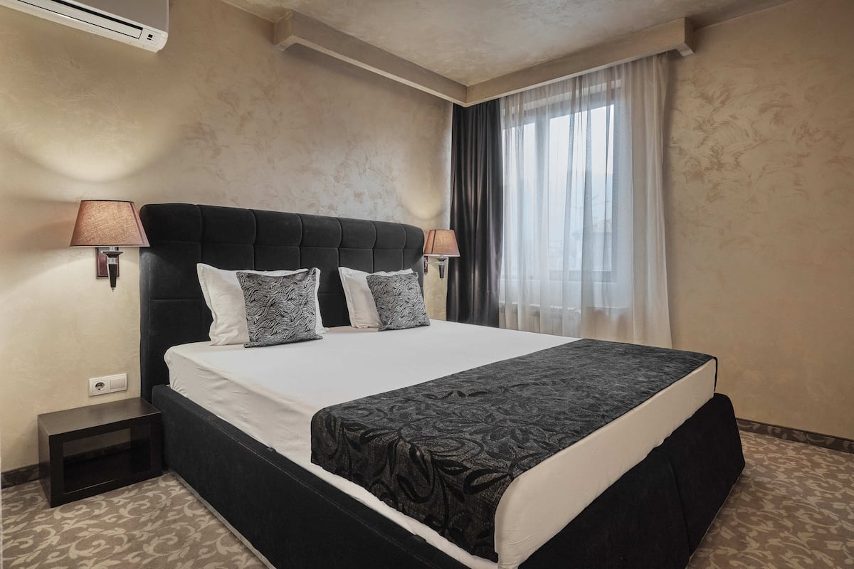 A neatly made bed with a black headboard, two bedside lamps, and a window with sheer curtains.