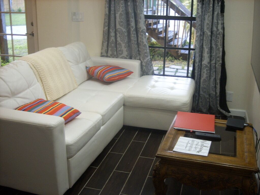 Un sofá de cuero blanco con almohadones de colores, una mesita de madera con una carpeta roja y una ventana con cortinas estampadas.