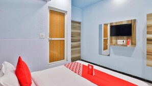 Ein Hotelzimmer mit einem Bett, einem roten Kissen, einem Teppich mit rotem und weißem Muster, einem an der Wand befestigten Fernseher und einer Holztür.