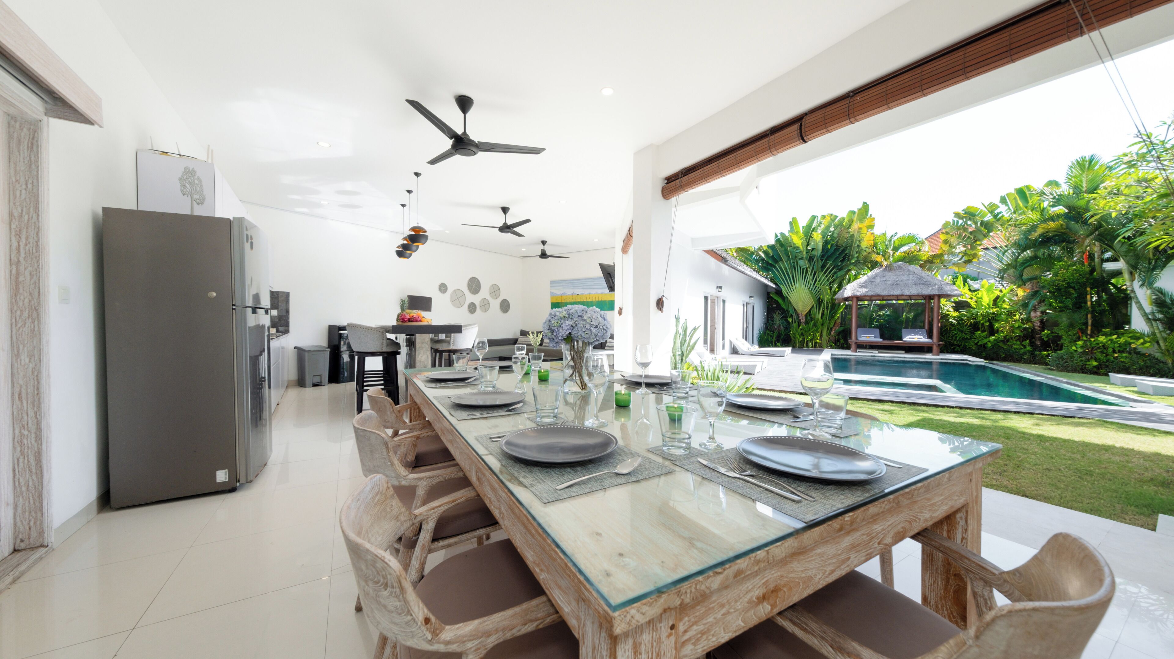 Une cuisine moderne dotée d’un grand îlot central, d’un coin repas avec une table en verre et d’une vue sur la piscine agrémentée de plantes tropicales.