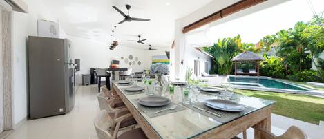 Une cuisine moderne dotée d’un grand îlot central, d’un coin repas avec une table en verre et d’une vue sur la piscine agrémentée de plantes tropicales.