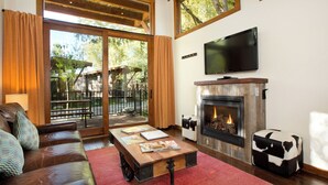 A living room with a leather sofa, a wooden coffee table, a fireplace, and a television mounted on the wall. - Cabin (Modern Rustic Wedge) (Wilson)