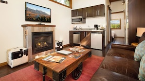 A cozy living room with a fireplace, a dining table with magazines, and a kitchen area with modern appliances. - Cabin (Modern Rustic Wedge) (Wilson)