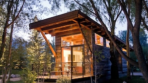 A log cabin with a wooden deck, large windows, and a stone foundation surrounded by trees. - Cabin (Modern Rustic Wedge) (Wilson)