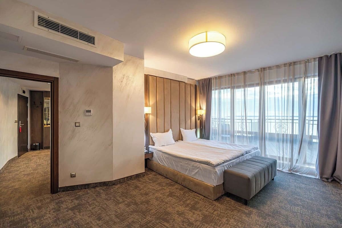Une chambre d’hôtel avec un grand lit, un banc, un radiateur fixé au mur, un luminaire au plafond et une vue sur l’extérieur.