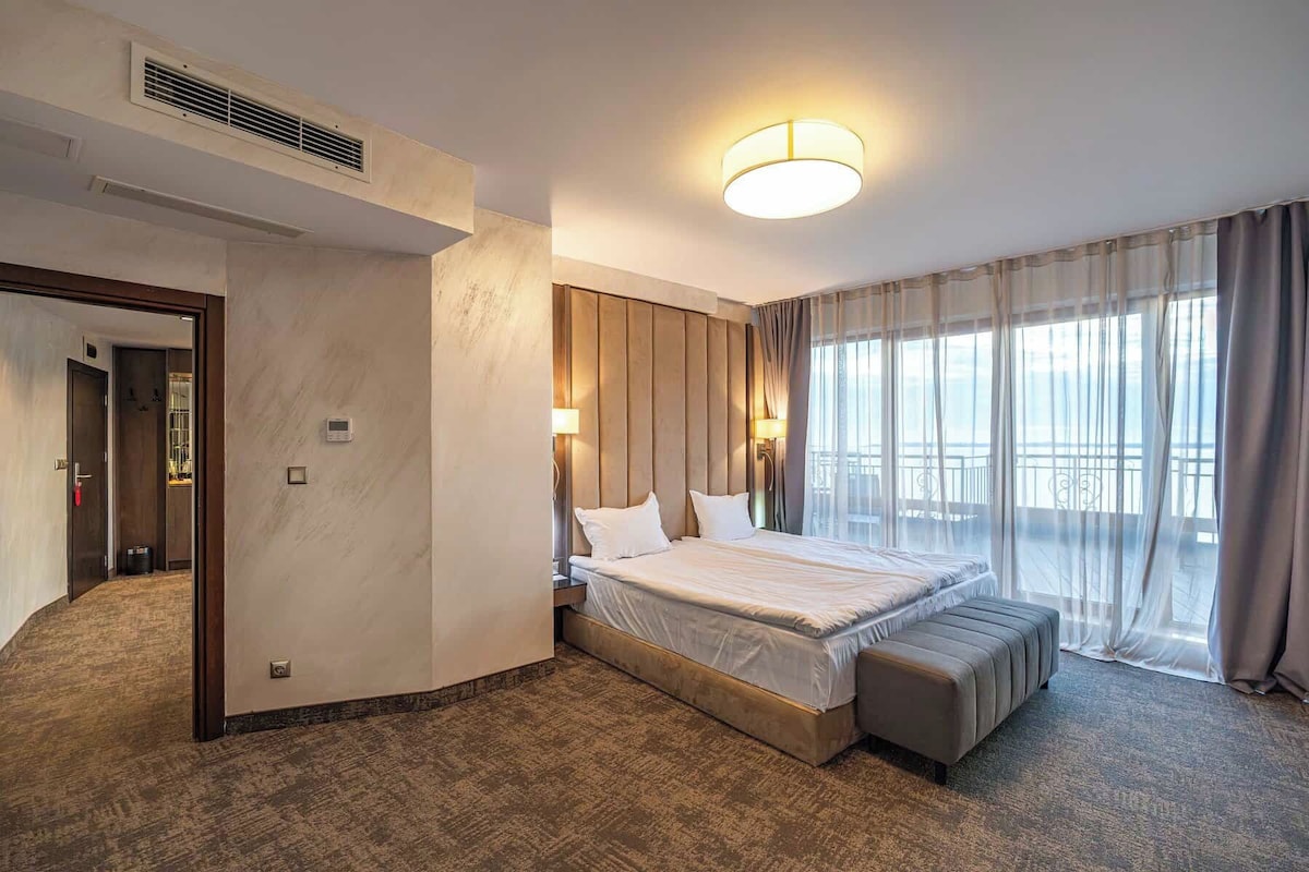 A hotel room with a large bed, a bench, a wall-mounted heater, a ceiling light, and a view of the outdoors.