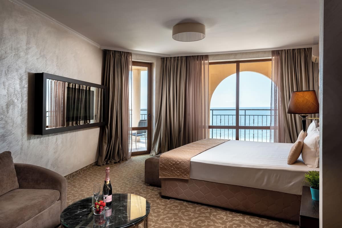 Une chambre d’hôtel avec un lit, un canapé, une petite table avec une bouteille et des verres, et une grande fenêtre avec des rideaux.