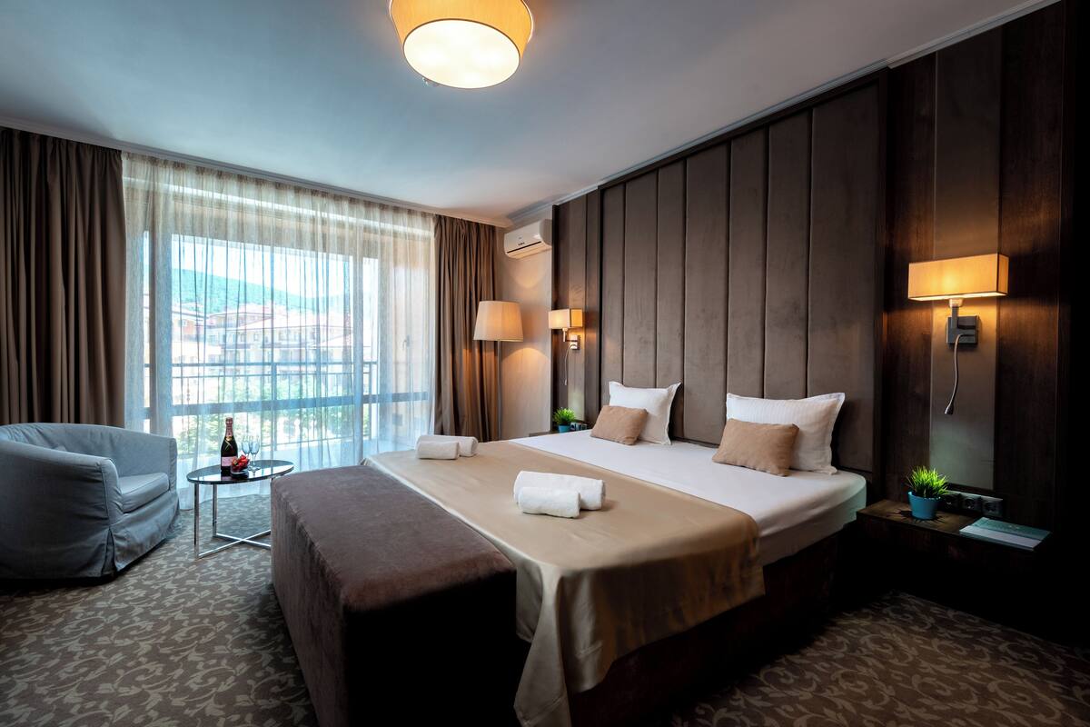 Une chambre d’hôtel avec un grand lit, un coin salon, une petite table avec une bouteille et des verres, et une vue sur les bâtiments par la fenêtre.