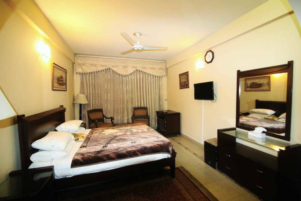 A hotel room with a bed, a ceiling fan, a TV, a mirror, and a clock on the wall.
