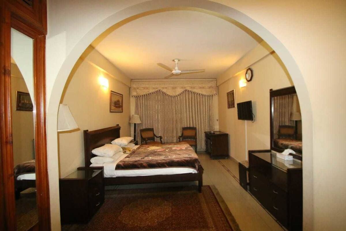 A hotel room with a bed, a desk, a chair, a TV, and a clock on the wall.