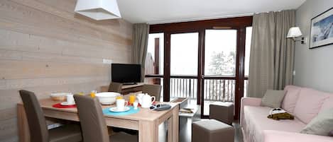 A room with a wooden wall, a dining table with chairs, a sofa, a TV, and a balcony with curtains.