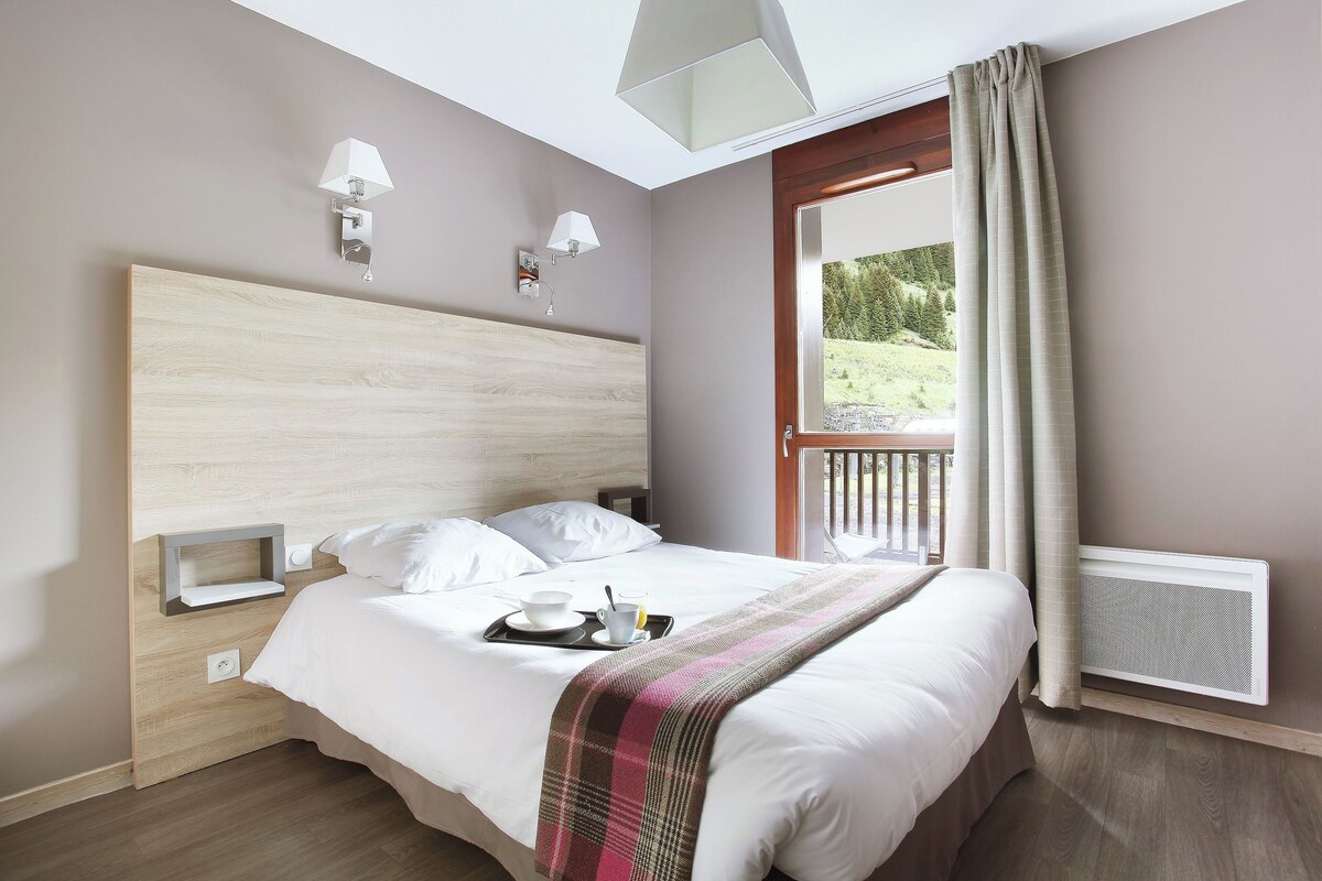A hotel room with a large bed, a nightstand, a wall-mounted lamp, a window with curtains, and a view of the outdoors.