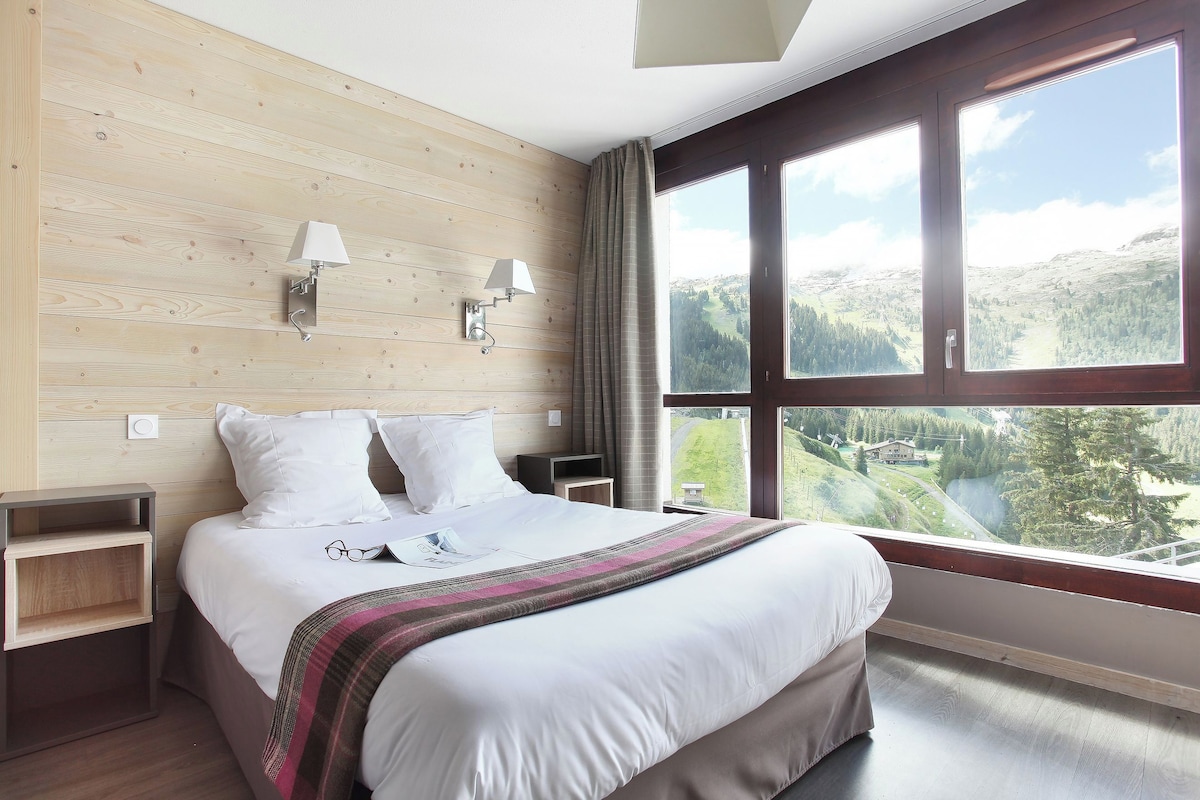 Une chambre d’hôtel avec un grand lit, deux tables de chevet, une fenêtre donnant sur un paysage montagneux et un lambris en bois.