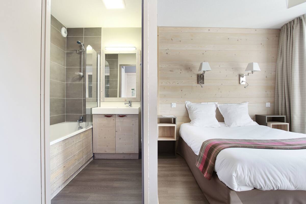 A modern hotel room with a shower, a wooden vanity, a bed with white linens, and a nightstand with a lamp.