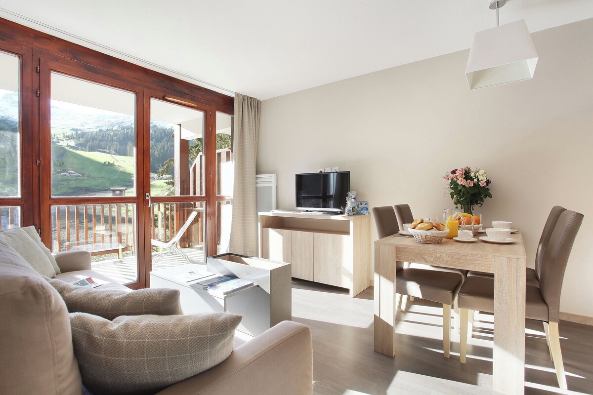 A modern living room with a sofa, a dining table with chairs, a TV, and a view of the outdoors.