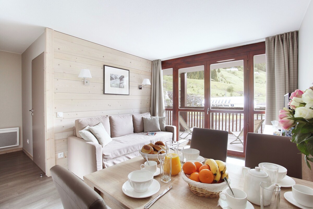 A cozy room with a wooden wall, a sofa, a dining table set with breakfast items, and a view of a garden through large windows.