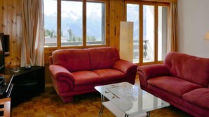A living room with two red sofas, a glass coffee table, and a TV on a wooden cabinet. Large windows offer a view of mountains. - Chaedoz 13 by Interhome (Nendaz)