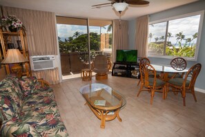 A living room with a sofa, a glass coffee table, wicker chairs, a television, and a balcony with a view of palm trees. - Nani Kai Hale #301 on Sugar Beach (Kihei)