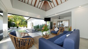A modern living room with a glass coffee table, blue sofas, and wicker chairs. - Villa Hiburan 2 by Villa Finder (Seminyak)
