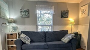 A living room with a grey sofa, a coffee table with a plate of oysters, a window with blinds, and wall art. - A Gnome Home: Convenient to Restaurants and Coffee Shops (Ocracoke)