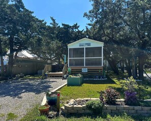 A small, single-story cabin with a porch, surrounded by trees and a gravel driveway.