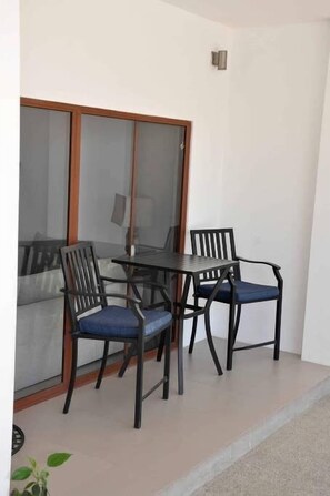 Uma varanda com mesa e duas cadeiras, uma porta de vidro deslizante e um pequeno vaso com planta.