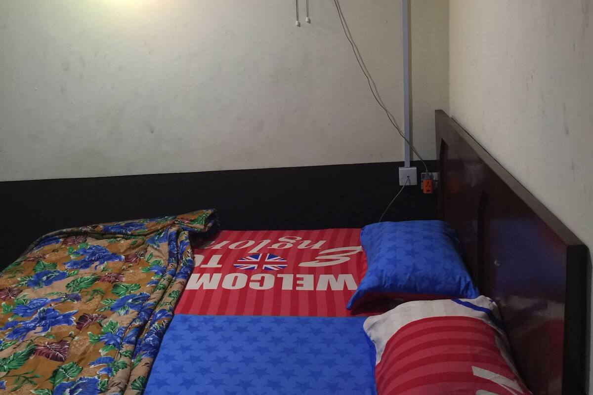 A single bed with a floral bedspread and a red and white striped pillow. There is a ceiling fan and an air conditioning unit mounted on the wall.
