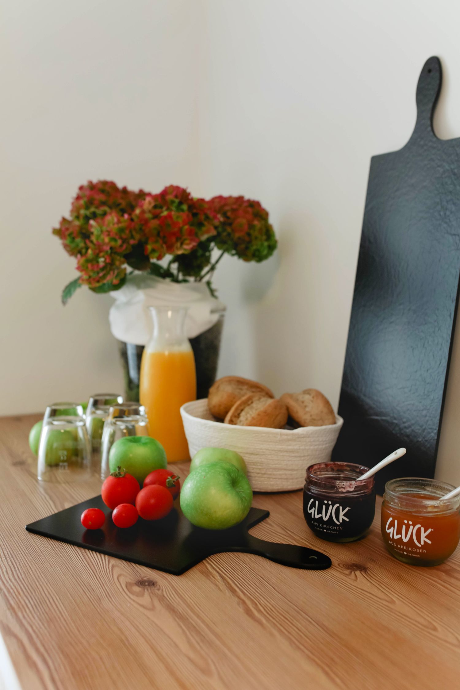 A wooden table with a bowl of apples, a cutting board, and jars of Gluck apricot jam.