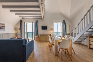 Ampla sala de estar com mesa de jantar, cadeiras e sofá. Escada à direita e varanda com cortinas.
