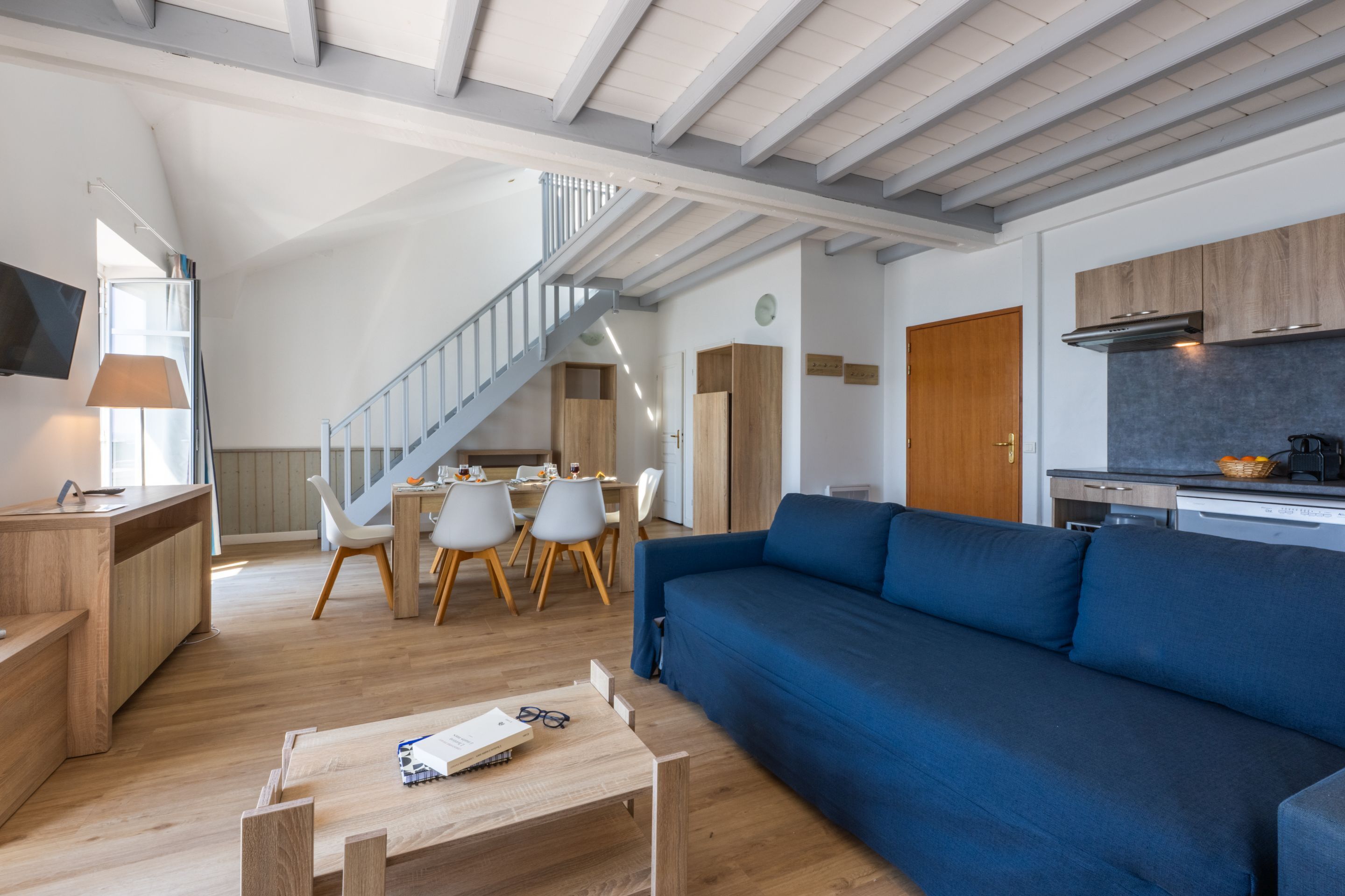 A modern living room with a blue sofa, wooden dining table, and chairs. A staircase leads to an upper level.