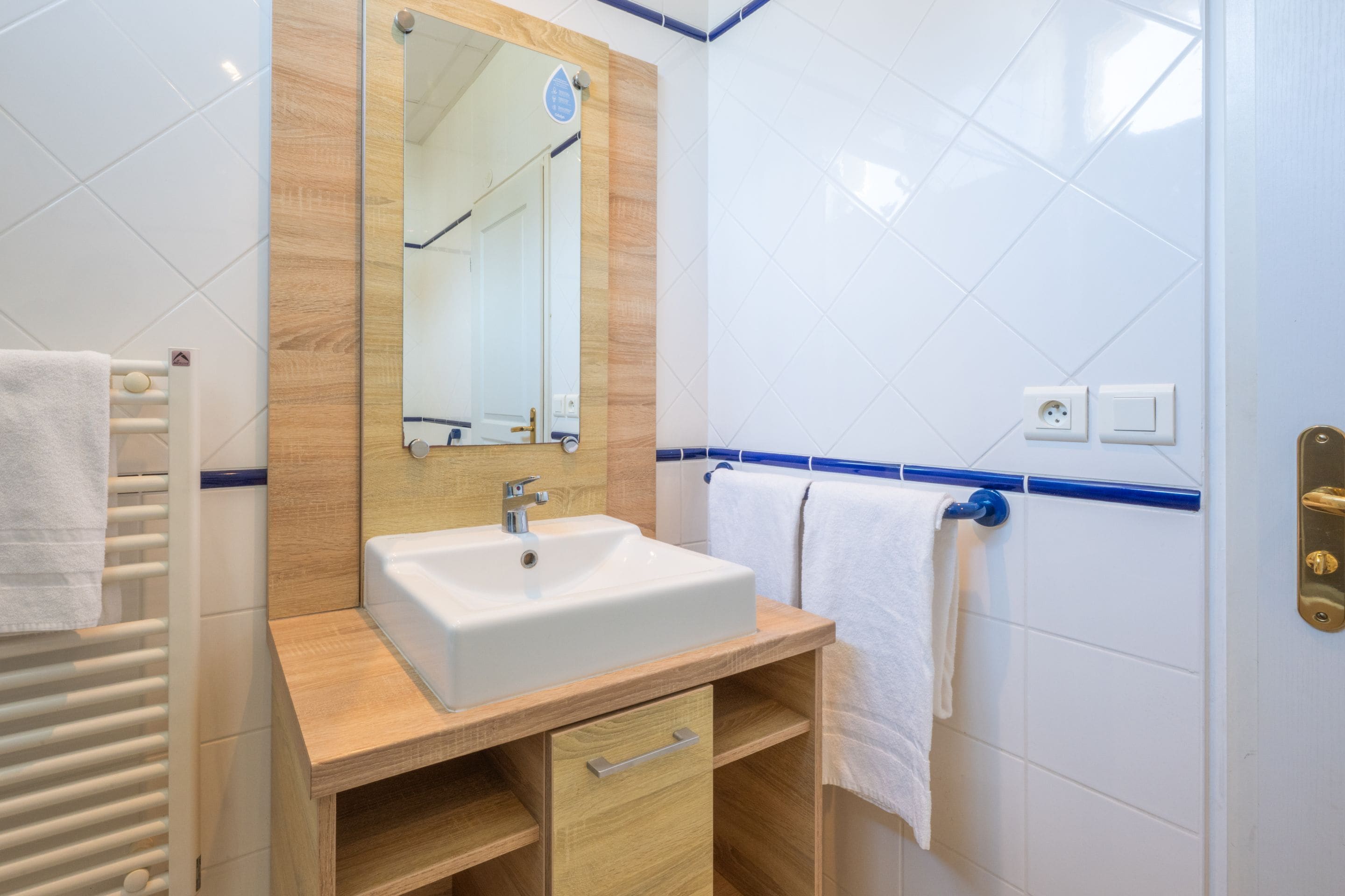 Un baño con lavamanos blanco, tocador de madera y espejo. Hay toallas colgadas en un perchero y un calentador de toallas.