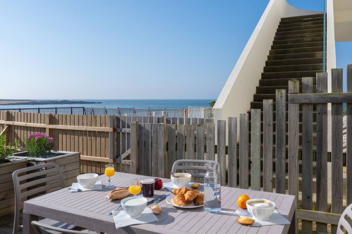 Une table de petit-déjeuner avec des croissants, du jus d’orange et du café, donnant sur la mer.
