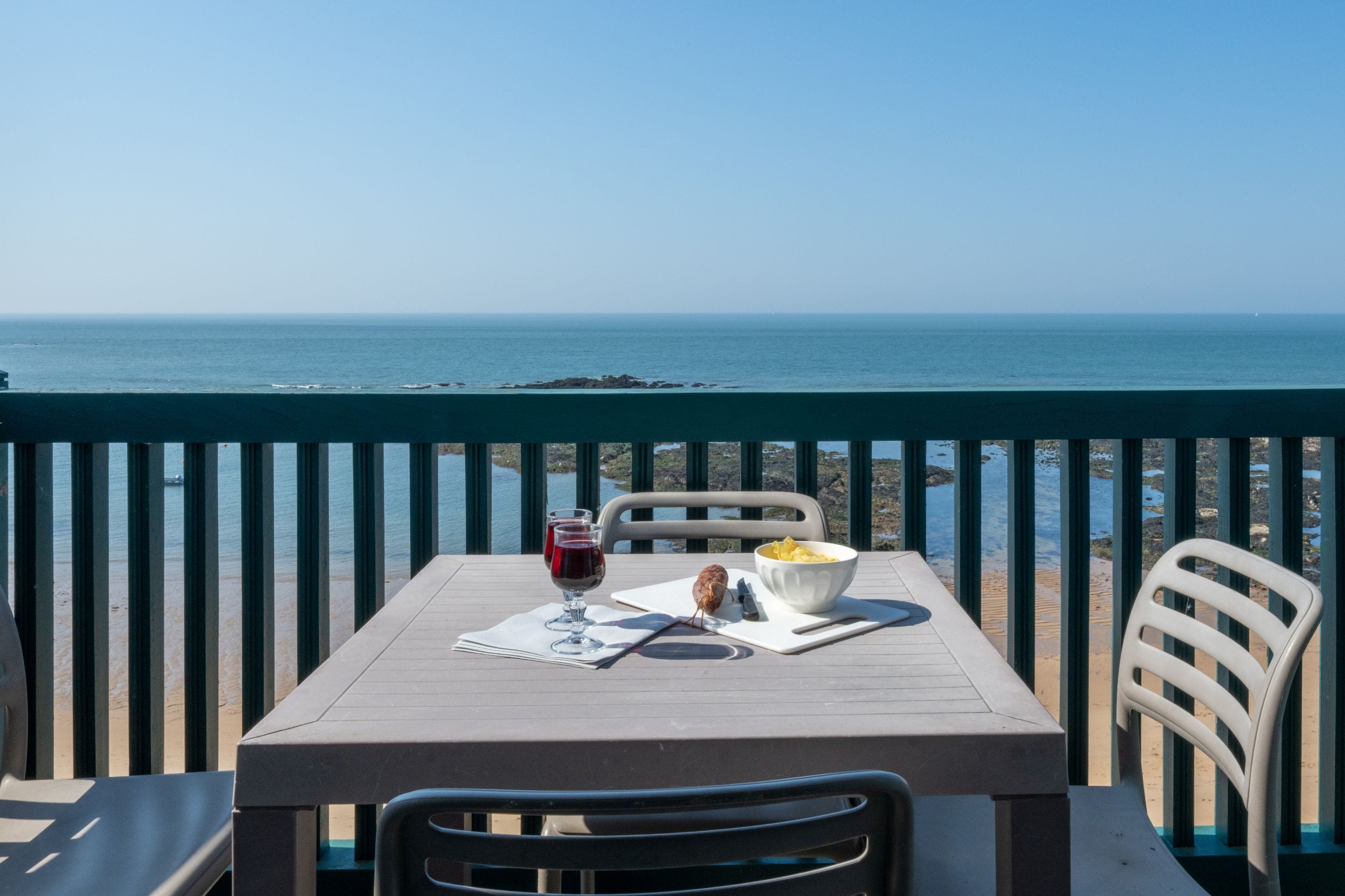 A table with a bowl of chips, a glass of red wine, and a menu on a balcony overlooking the sea.