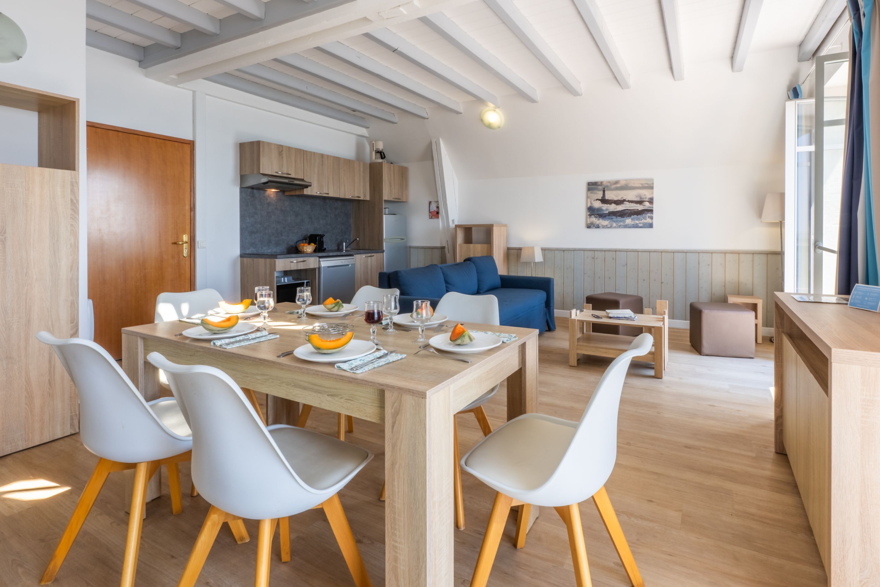 A modern kitchen with wooden cabinets, a dining table with chairs, and a blue sofa in a living area.