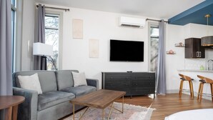 Un salon moderne avec un canapé gris, une table basse en bois, un téléviseur à écran plat, une commode de couleur foncée et un coin cuisine avec des tabourets de bar.