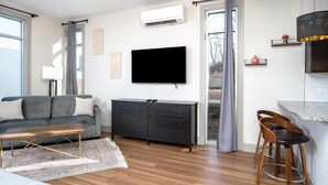 Un salon moderne comprenant un canapé, un téléviseur, une table basse en bois, une commode sombre et un coin cuisine avec un tabouret de bar.