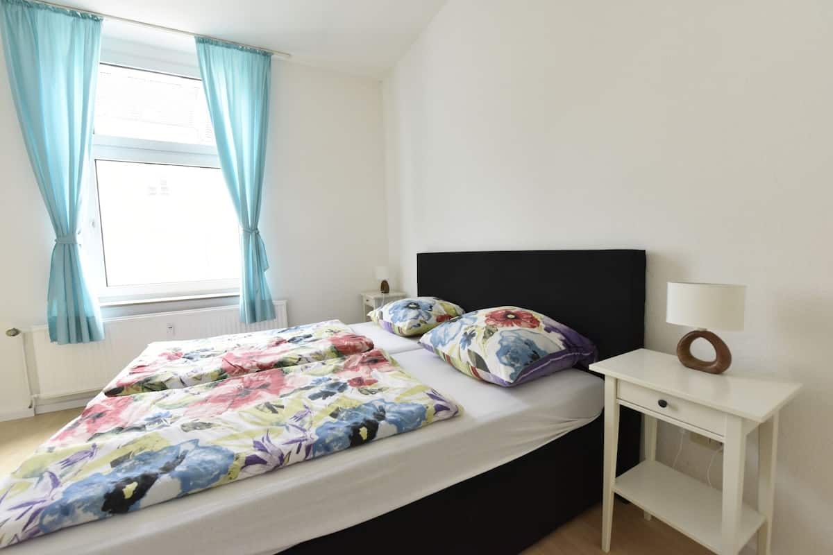 A bedroom with a bed, a bedside table, a lamp, and a window with curtains.