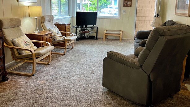 A living room with a beige carpet, a television, a wooden chair, a sofa, and a small table with a lamp.