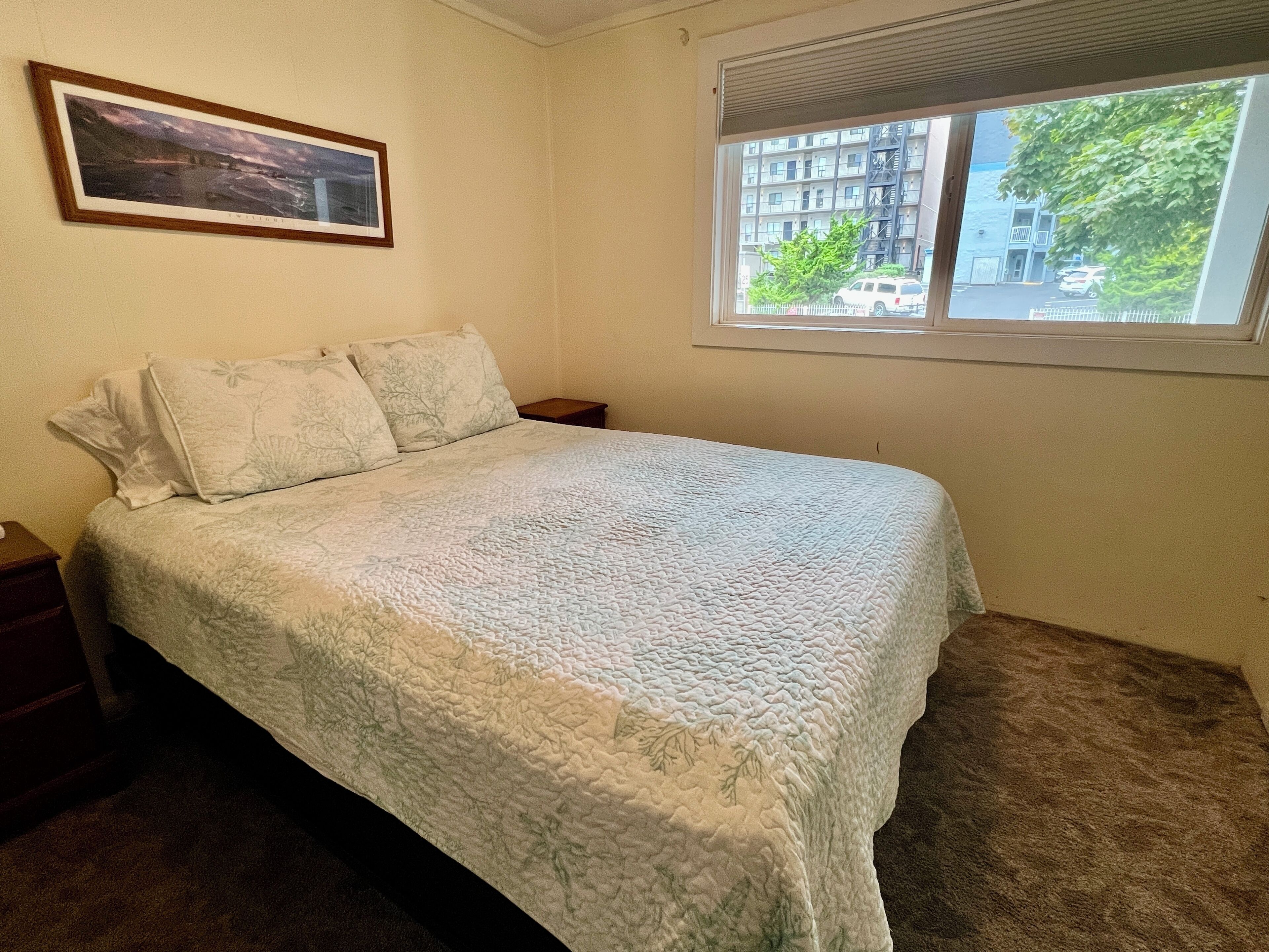 A bedroom with a bed, a nightstand, a window with a view of buildings and trees, and a framed picture on the wall.