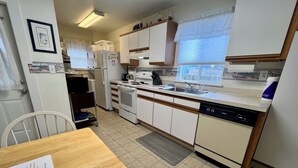 A compact kitchen with white cabinets, a dishwasher, a stove, and a sink. There is a window with blinds, a door, and a chair.