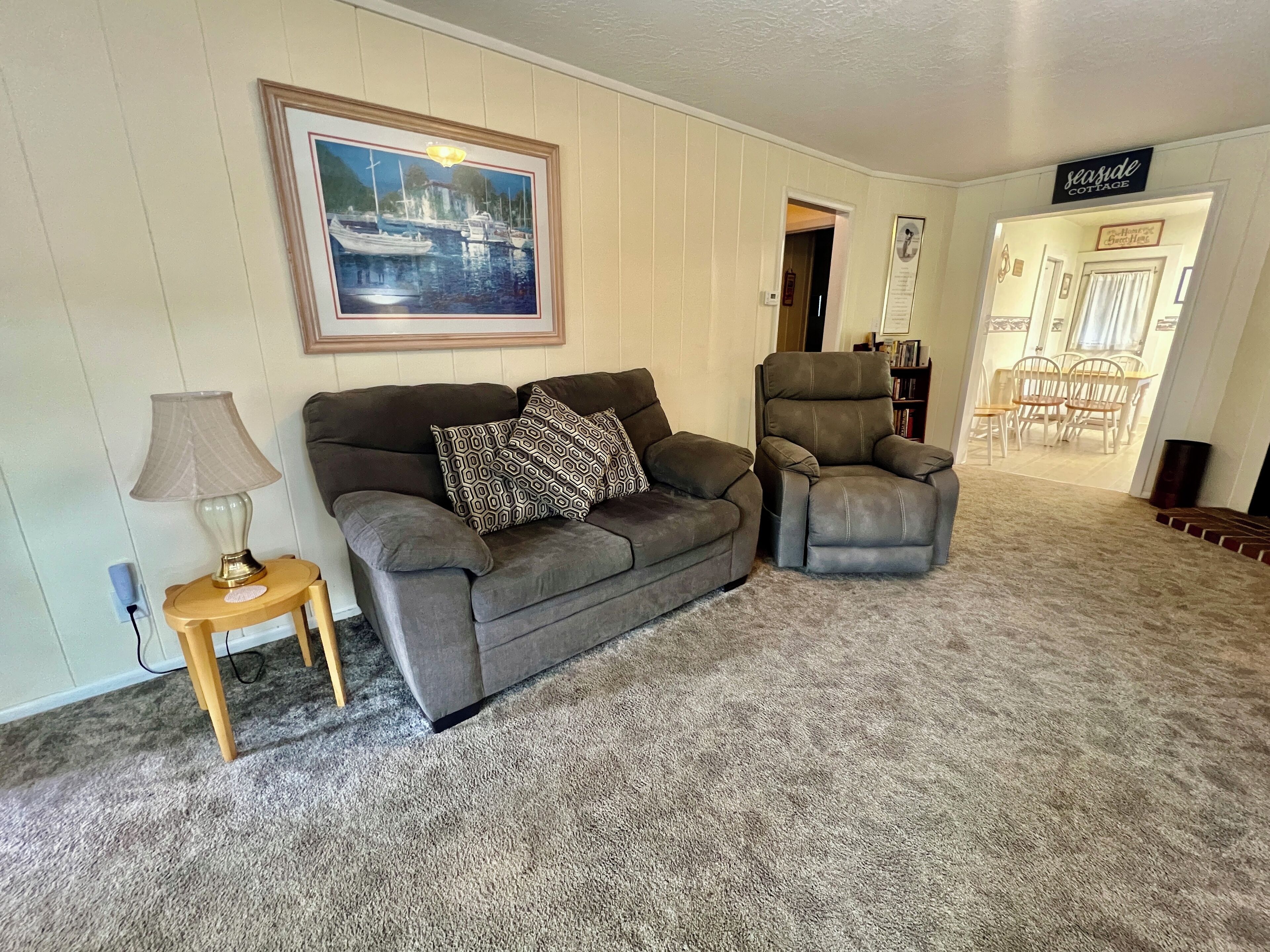 Un salon avec un tapis beige, un canapé gris agrémenté de coussins à motifs, une table d’appoint en bois avec une lampe, un fauteuil inclinable gris et un tableau encadré représentant un paysage portuaire.