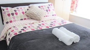 A bed with a dark grey bedspread, a pink and purple heart-patterned duvet, and a rolled white towel.
