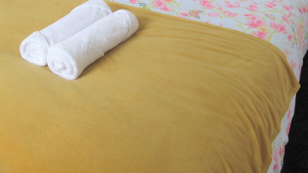 A neatly made bed with floral bedding, a yellow throw blanket, and two rolled towels.