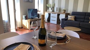 Zona pranzo con tavolo di legno apparecchiato per due, una bottiglia di vino e due bicchieri da vino. Sullo sfondo, un divano e una televisione.