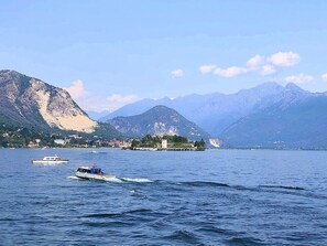 Um lago com barcos, uma pequena ilha com edifícios e montanhas ao fundo.