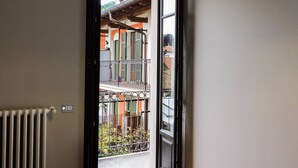 Una stanza con un termosifone, un letto con copripiumino decorato e una porta aperta che dà su un balcone con vista su un edificio e del verde.