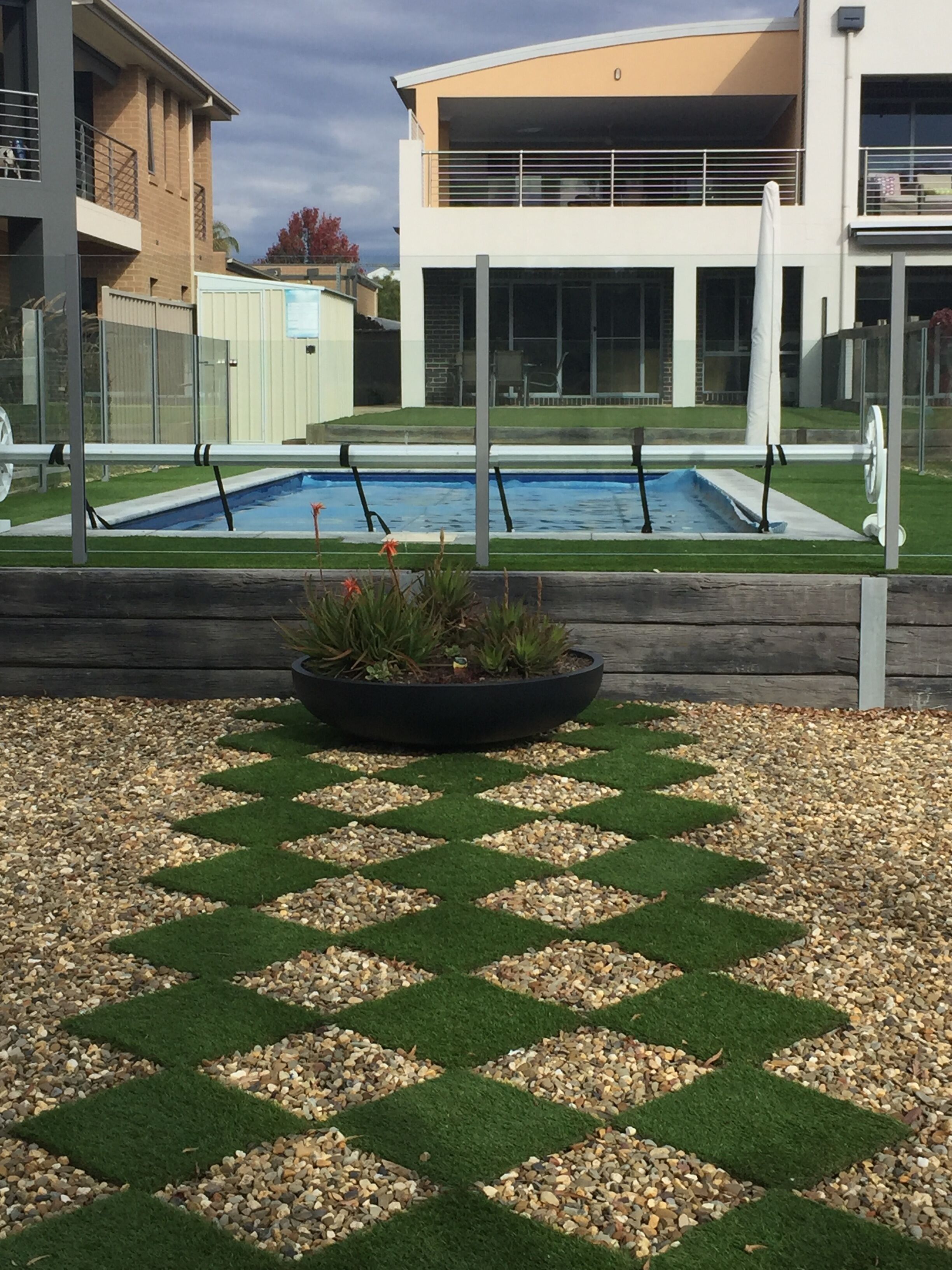 A swimming pool with a geometric pattern of artificial grass and pebbles, surrounded by a building with a modern design.