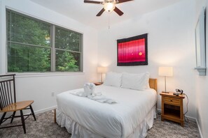 A bedroom with a large window, a ceiling fan, a bed with white bedding, two bedside tables with lamps, and a chair. - Sleepy Hollow Beach Resort introduces Patio 33 (South Haven)