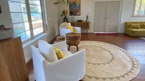 A spacious living room with a large window, a wooden floor, a white sofa, a yellow armchair, and a round rug. - Large villa Le Ficus by the sea with swimming pool (PAEA)