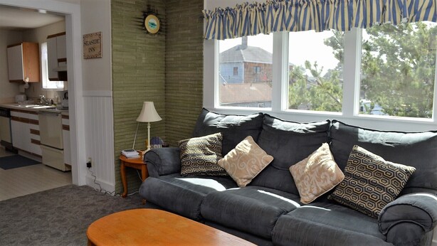 Een woonkamer met een grijze bank, gekleurde kussens, een houten bijzettafeltje, een lamp en uitzicht op een huis door een raam.